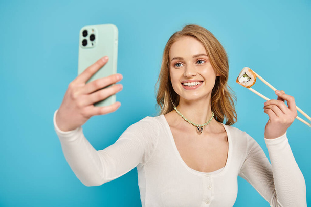 Een stijlvolle vrouw met blond haar met sushi en eetstokjes in de hand en een mobiele telefoon in de andere. - Foto, afbeelding