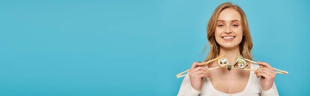 Μια λαμπερή γυναίκα με ξανθά μαλλιά που κρατάει δύο ξυλάκια με σούσι σκαρφαλωμένο πάνω τους μπροστά στο πρόσωπό της.. - Φωτογραφία, εικόνα