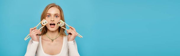 A beautiful woman with blonde hair holds up chopsticks with sushi near her face - Photo, Image