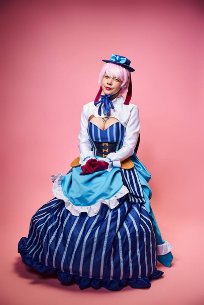 seductive sexy female cosplayer in blue hat and vibrant attire looking at camera on pink backdrop - Foto, afbeelding