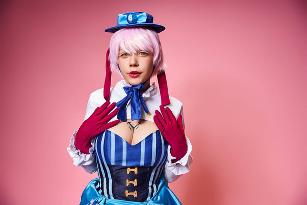 beautiful stylish female cosplayer in blue hat and vivid dress looking at camera on pink backdrop - Фото, зображення