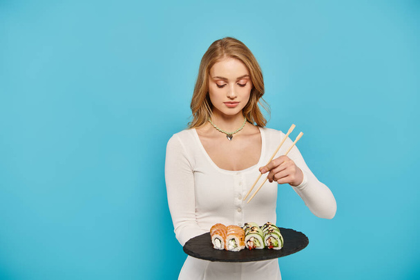 Piękna blondynka delikatnie trzyma talerz sushi i pałeczek, pokazując sztukę korzystania z kuchni azjatyckiej. - Zdjęcie, obraz