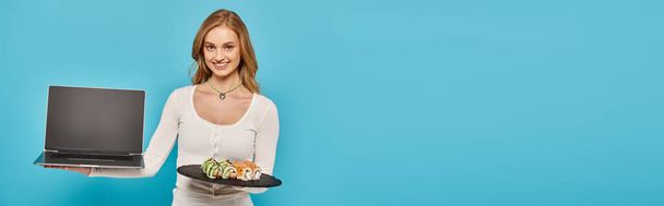 A blonde woman balances sushi on a plate while holding laptop, embodying a modern foodie lifestyle. - Photo, Image