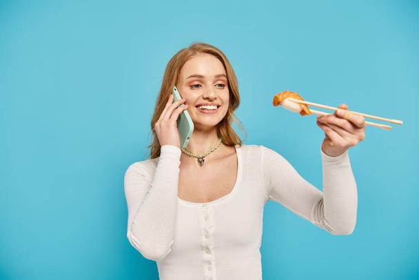Eine blonde Frau hält zart Essstäbchen in der Hand, während sie am Telefon plaudert und asiatische Küche genießt. - Foto, Bild