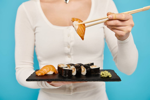 Ausgeschnittene Ansicht der Frau hält elegant einen Teller mit Sushi und Essstäbchen in der Hand und genießt jeden Bissen mit gelassener Miene. - Foto, Bild