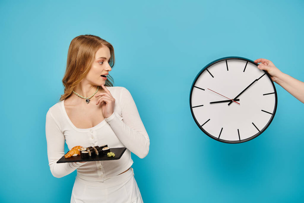 Uma mulher com cabelo loiro segurando um prato de comida asiática na frente de um relógio, mostrando uma hora de refeição tentadora. - Foto, Imagem