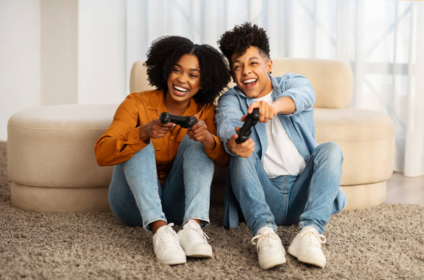 Due gioiosi giovani afroamericani seduti sul tappeto in un soggiorno, ridono e si divertono mentre giocano ai videogiochi insieme in un ambiente familiare rilassato - Foto, immagini