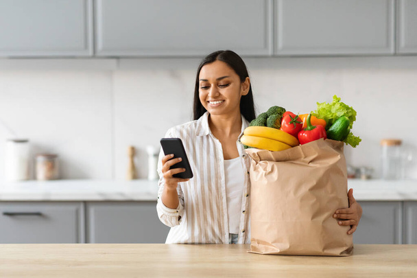 Fröhliche junge indische Frau bestellt Lebensmittel online, steht neben dem Tisch in der Küche, hält Papiertüten voller frischem Bio-Obst und -Gemüse in der Hand, per Smartphone, Handy-App, kopiert Leerstellen - Foto, Bild