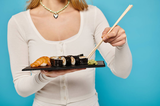 Vista recortada de la mujer con estilo sostiene un plato de sushi y palillos, listo para disfrutar de la deliciosa delicia asiática. - Foto, imagen