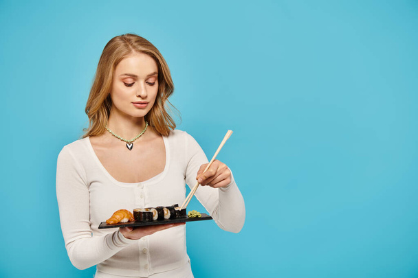 Una mujer rubia con gracia sostiene un plato de sushi con palillos, mostrando la belleza de la cocina asiática. - Foto, imagen