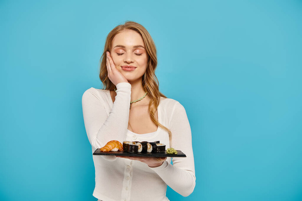 A blonde woman gracefully holds a tray of sushi against a serene blue background. - Photo, Image