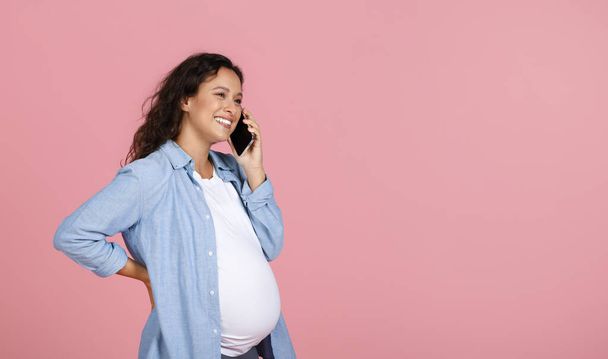 Šťastná mladá těhotná žena na sobě ležérní oblečení mít telefonní rozhovor s přítelem, při pohledu na kopírovací prostor pro reklamu. Čekající dáma volá svého lékaře, růžové pozadí - Fotografie, Obrázek