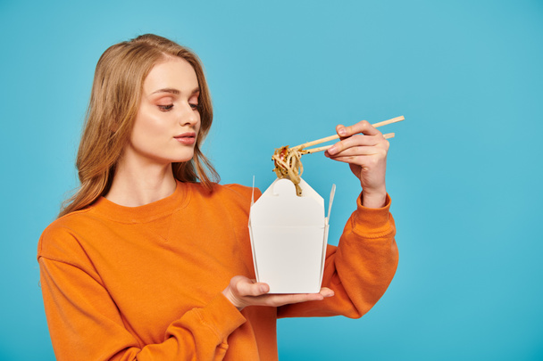 Блондинка в оранжевом свитере держит белый контейнер с лапшой, демонстрируя азиатскую кухню палочками для еды. - Фото, изображение