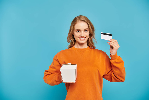 Μια κομψή γυναίκα εμφανίζεται ικανοποιημένη, καθώς κρατά μια πιστωτική κάρτα στο ένα χέρι και ένα κουτί τροφίμων στο άλλο, συμβολίζοντας online αγορές. - Φωτογραφία, εικόνα