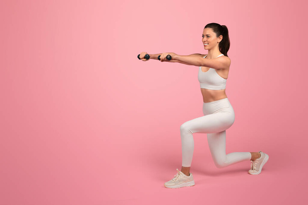 Щаслива сильна тисячолітня біла жінка в білому спортивному одязі, присідає гантелями, насолоджується тренуванням, активним способом життя, ізольована на рожевому тлі. Охорона здоров'я, спорт і втрата ваги - Фото, зображення