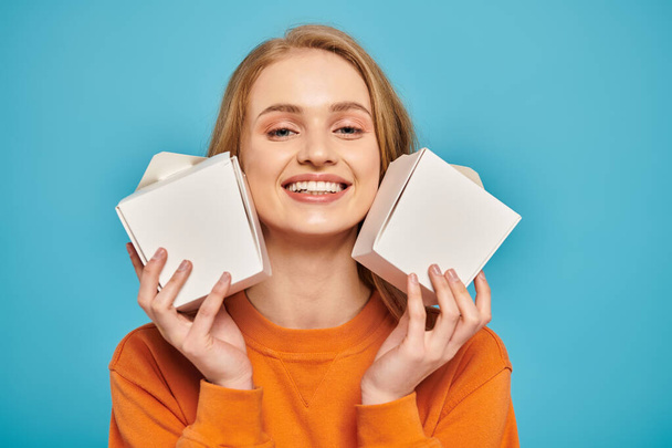 A beautiful blonde woman holds two food boxes in front of her face, concealing her expression with intrigue. - Photo, Image