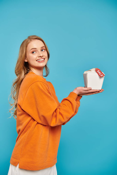 Μια κομψή ξανθιά γυναίκα με χάρη κρατά κουτί τροφίμων με ετικέτα πώλησης, αποπνέοντας κομψότητα και κομψότητα. - Φωτογραφία, εικόνα
