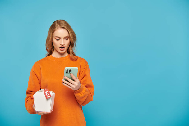 オレンジ色のセーターのスタイリッシュな女性は自信を持って携帯電話を保持し,その上に食べ物のセールスタグ. - 写真・画像
