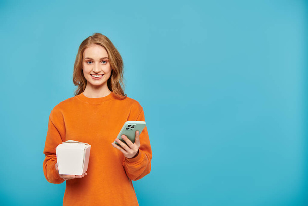 Una donna con i capelli biondi che tiene la scatola del cibo e un telefono cellulare mentre sullo sfondo blu - Foto, immagini