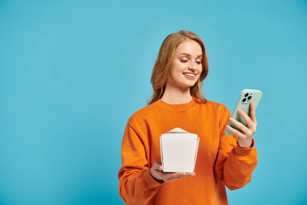 Une femme blonde équilibre sans effort une boîte de nourriture asiatique dans une main tout en faisant défiler son téléphone portable avec l'autre. - Photo, image