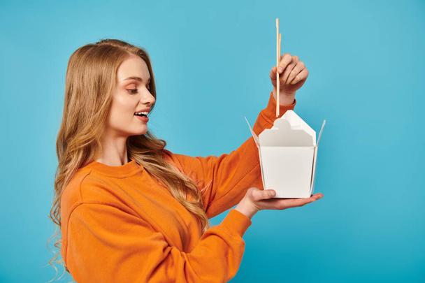A stylish woman in an orange shirt confidently holds a box with Asian food and chopsticks. - Valokuva, kuva