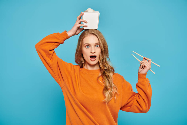 Una bella donna bionda in un maglione arancione tiene giocosamente le bacchette sopra la testa con una scatola di cibo nelle vicinanze. - Foto, immagini