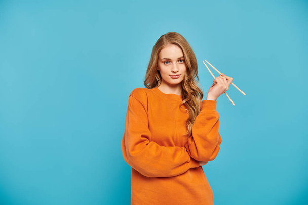 A beautiful woman in an orange sweater delicately holds a pair of chopsticks, ready to enjoy a delicious Asian meal. - Photo, Image