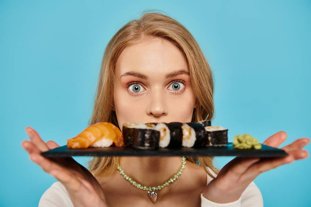A beautiful blonde woman gracefully holds a plate filled with a variety of sushi, showcasing the delicious Asian cuisine. - Fotografie, Obrázek