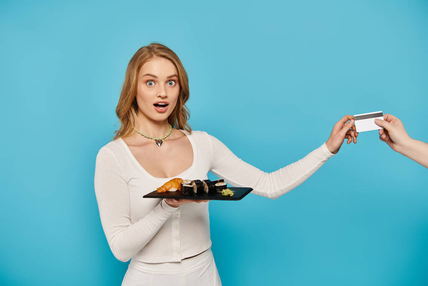 Eine schöne blonde Frau in einem weißen Kleid hält elegant ein Tablett mit köstlicher asiatischer Küche in der Hand, Hand mit Kreditkarte. - Foto, Bild