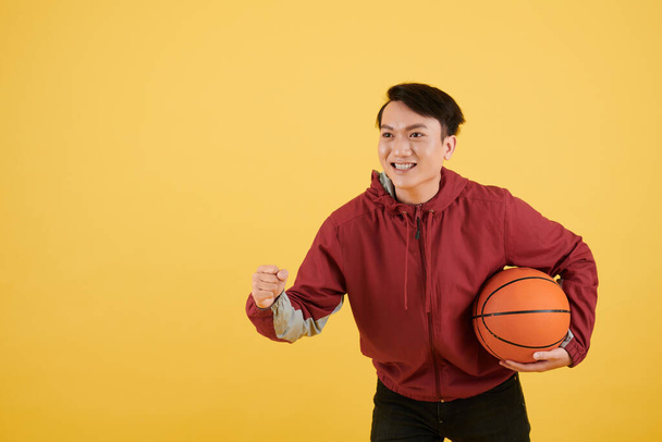 Basketbol topuyla favori takımı için tezahürat yapan heyecanlı Vietnamlı genç adamın portresi. - Fotoğraf, Görsel
