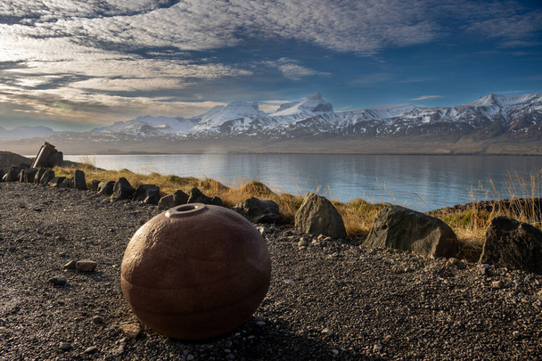 Ржавый большой железный шар на побережье фьорда. Горы, покрытые снегом с другой стороны. Голубое небо с белыми облаками. Баккагерди, Северо-Восточная Исландия. - Фото, изображение