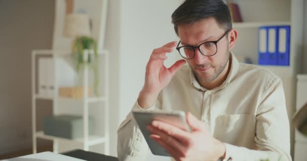 Nuori liikemies, joka säätää silmälaseja ja käyttää digitaalista tablettia istuessaan toimistossa - Materiaali, video