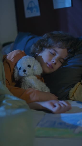 Beyaz tenli bir çocuğun yatakta uyuduğu, geceleri doldurulmuş beyaz bir köpek oyuncağına sarıldığının dikey el kamerası görüntüleri. - Video, Çekim