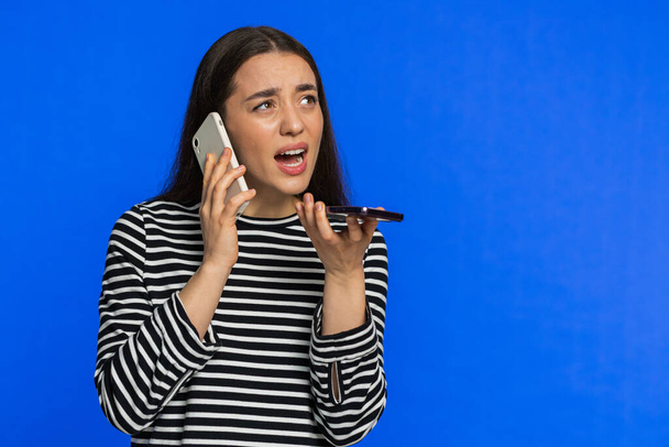 Ενοχλημένη αγχωμένη γυναίκα που μιλάει ουρλιάζοντας σε δύο κινητά τηλέφωνα και συζητάει διαφορές παραπόνων για διαμάχες για διαμάχες για επίλυση προβλημάτων. Νεαρή κοπέλα απομονωμένη σε μπλε φόντο. Αντιγραφή χώρου - Φωτογραφία, εικόνα