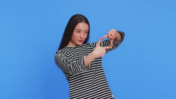 Femme caucasienne photographe gestuelle cadre photo avec les mains, regarde à travers les doigts et en se concentrant sur moment intéressant imitant zoom et recadrage belle image. Jeune fille isolée sur fond bleu - Séquence, vidéo