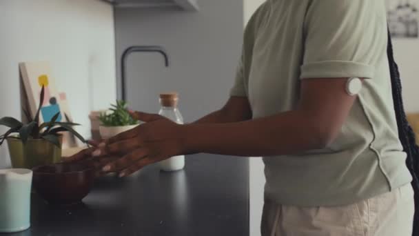 Medium shot of midsection of unrecognizable black woman with glucose monitor patch on arm and long braids walking up to kitchen table and pouring milk into bowl for breakfast - Footage, Video