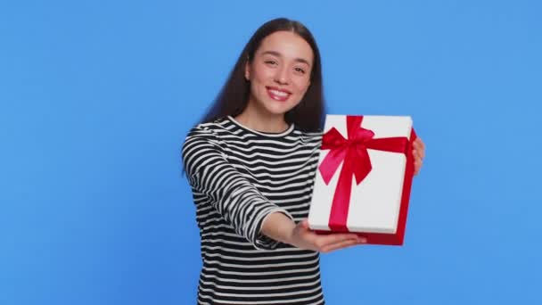 Mujer caucásica sonriente positiva que presenta caja de regalo de cumpleaños se extiende las manos ofrecen regalo envuelto bono de carrera que celebra la venta de descuento de promoción de fiesta. Chica bonita aislada sobre fondo azul - Imágenes, Vídeo