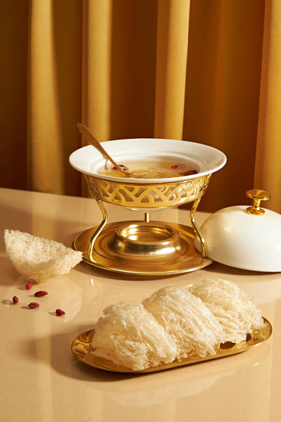 Vogelnestsuppe, die in einem Futterwärmer mit einem Löffel enthalten ist, eine goldene Schüssel essbaren Vogelnestes, die mit getrockneten Goji-Beeren auf einem Spiegeltisch ausgestellt ist. Vogelnest ist kostbar und in der Natur zu finden - Foto, Bild