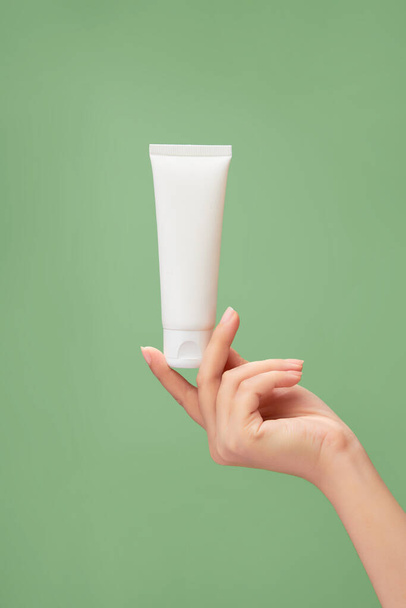 Egy pasztell zöld háttér, női kéz modell kezében egy fehér cső címke nélkül. Szépségápolási termék mockup. Természetes bőrápoló termékek koncepciója - Fotó, kép