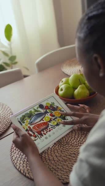 Середній вертикальний задній знімок молодої чорної жінки з діабетом, плетене волосся сидить за кухонним столом вдома, дивлячись на картину зі здоровою діабетичною їжею на планшетному комп'ютері при вивченні харчування - Кадри, відео