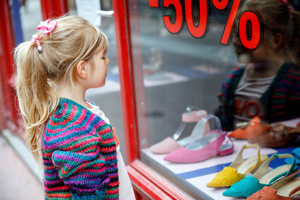 Pikkutyttö katsoo ikkunaa lähellä ostoskeskusta. Suloinen onnellinen lapsi katselee kenkiä varastossa ikkunassa - Valokuva, kuva