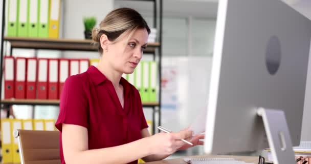 Mujer de negocios seriamente enfocada está escribiendo en la computadora y sosteniendo documentos en las manos. Empresaria prepara informe con un análisis de los resultados del trabajo - Imágenes, Vídeo
