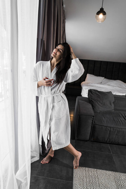 Egy kedves nő felébred egy hotelszobában, és élvezi a kilátást az ablakból télen. Kávét az ágyban. Szállodai üdülési koncepció - Fotó, kép