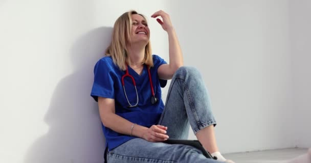 Κουρασμένος χειρουργός κάθεται στο πάτωμα στο λόμπι της κλινικής γελώντας υστερικά. Γυναίκα γιατρός και νευρική ένταση στην εργασία - Πλάνα, βίντεο