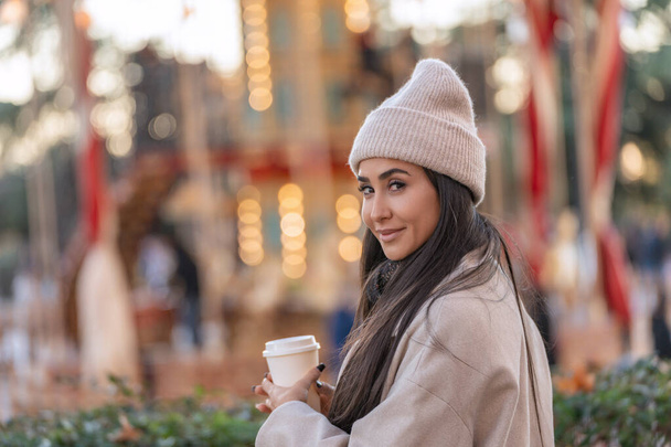 Parlak ışıklarla dolu bir atlıkarıncanın önünde elinde kahve fincanı tutan gülümseyen genç kadın..  - Fotoğraf, Görsel