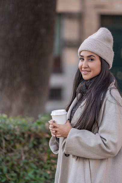 Elinde kahve fincanı olan şık genç bir kadın, kışa uygun giyinmiş, bir şehir caddesinde gülümsüyor..  - Fotoğraf, Görsel