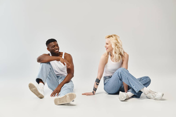 Ein junges multikulturelles Paar sitzt friedlich und verbunden auf dem Boden in einem Studio vor grauem Hintergrund. - Foto, Bild