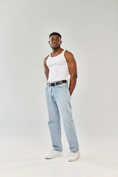 Nuori ja seksikäs Afrikkalainen Amerikkalainen mies luottavaisesti poseeraa valkoinen toppi ja farkut vastaan harmaa studio tausta. - Valokuva, kuva