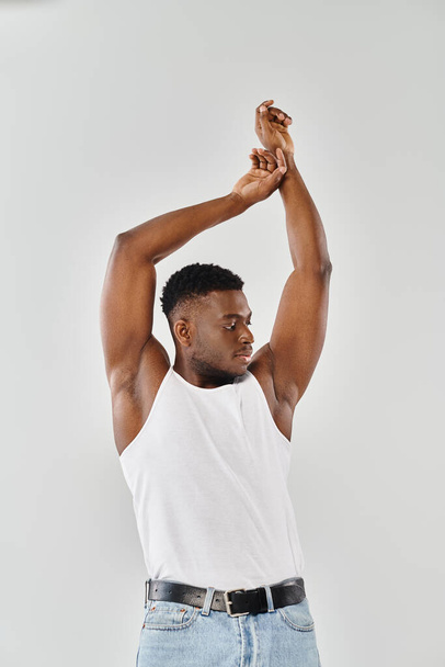 Un giovane afroamericano in canottiera bianca, che alza le mani in aria in uno studio su uno sfondo grigio. - Foto, immagini
