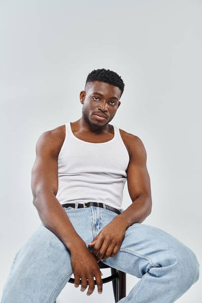 A young man of an interracial couple sits confidently atop a chair while wearing jeans on a grey studio background. - Photo, Image